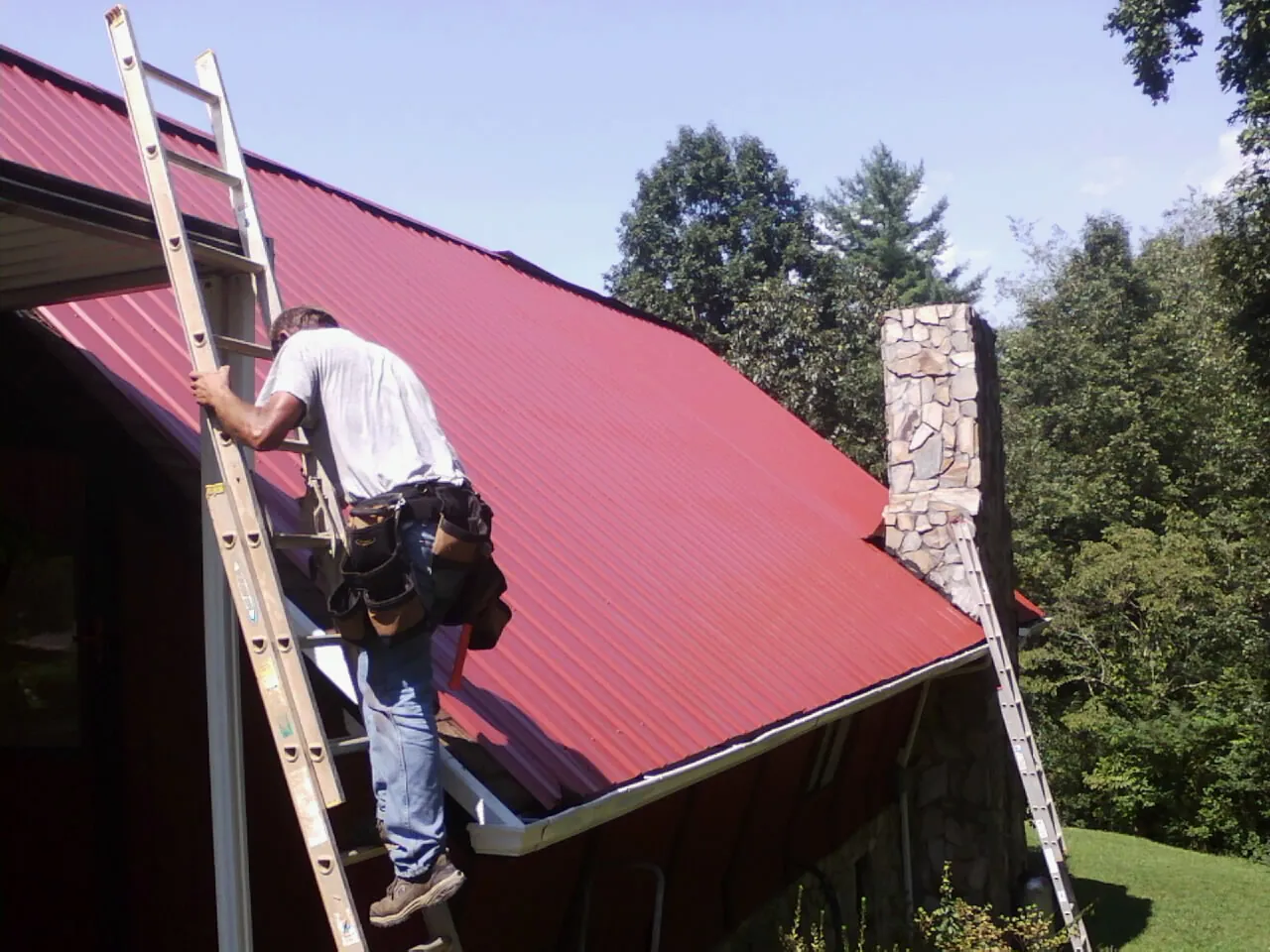 Roofer on a ladder beside a red metal roof and stone chimney — Soffit & Fascia Repair by Roofing Brothers in Bringhurst